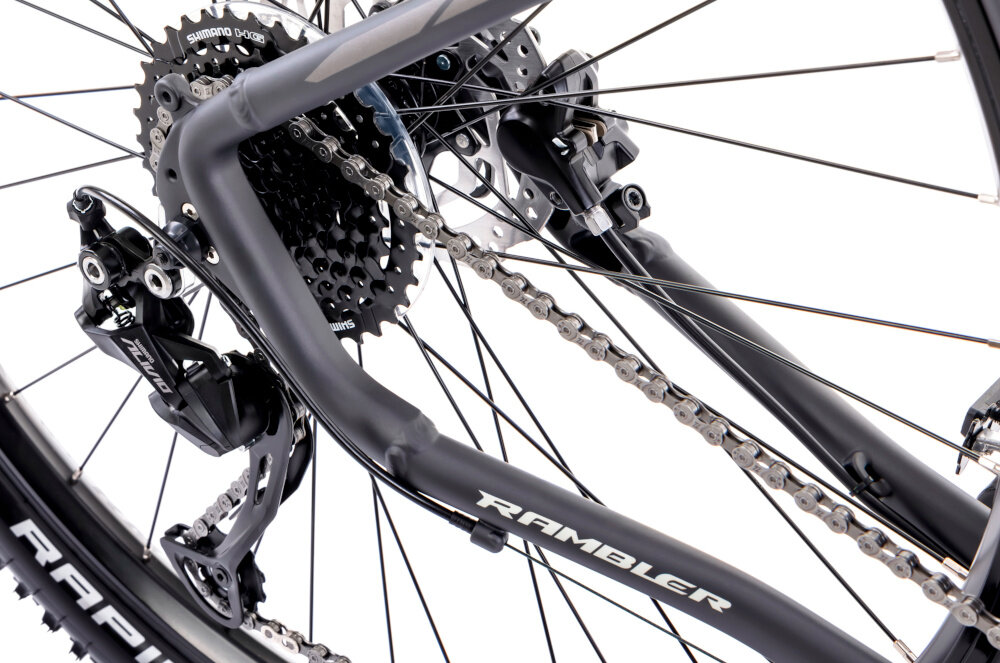 Zbliżenie na mechanizm napędowy roweru górskiego z widocznym napisem \'RAMBLER\' na ramie. Widać łańcuch, zębatki oraz przerzutkę marki Shimano.