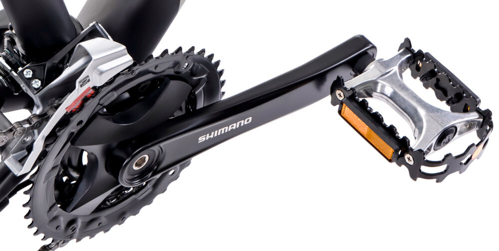 Zbliżenie na mechanizm korbowy roweru z widocznym napisem \'SHIMANO\' na ramieniu korby. Obok znajduje się pedał z metalowymi elementami i odblaskiem.