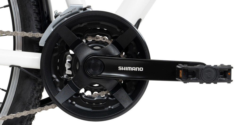 Zbliżenie na mechanizm korbowy roweru z widocznym napisem \'SHIMANO\' na ramieniu korby. W tle widać fragment białej ramy i opony roweru.