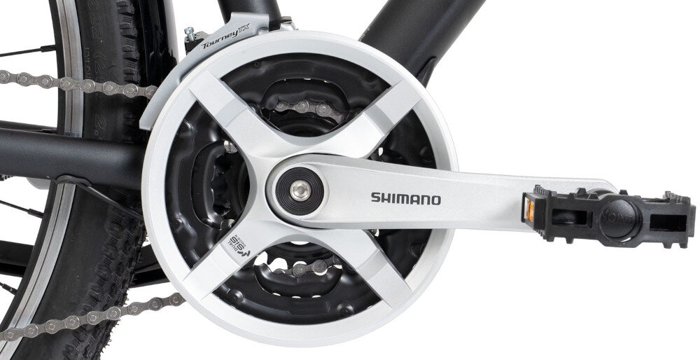 Zdjęcie przedstawia mechanizm korbowy roweru z widocznym napisem \'SHIMANO\' na ramieniu korby. W tle widać fragment opony i łańcucha rowerowego.