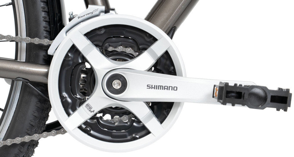 Zdjęcie przedstawia mechanizm korbowy roweru z widocznym napisem \'SHIMANO\' na ramieniu korby. W tle widać fragment opony i łańcucha rowerowego.