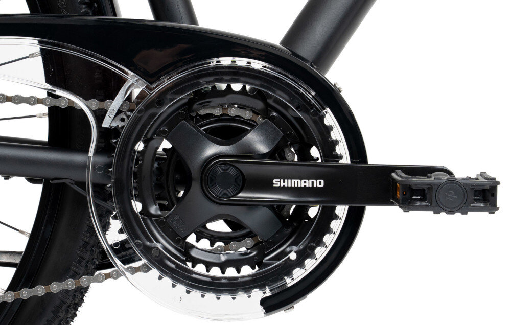 Zdjęcie przedstawia mechanizm korbowy roweru z widocznym napisem \'SHIMANO\' na ramieniu korby. Elementy są w kolorze czarnym z przezroczystą osłoną na łańcuch.