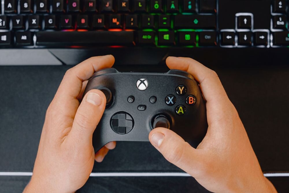 Osoba trzyma czarny kontroler do gier z widocznym logo Xbox. W tle znajduje się klawiatura z podświetleniem RGB.