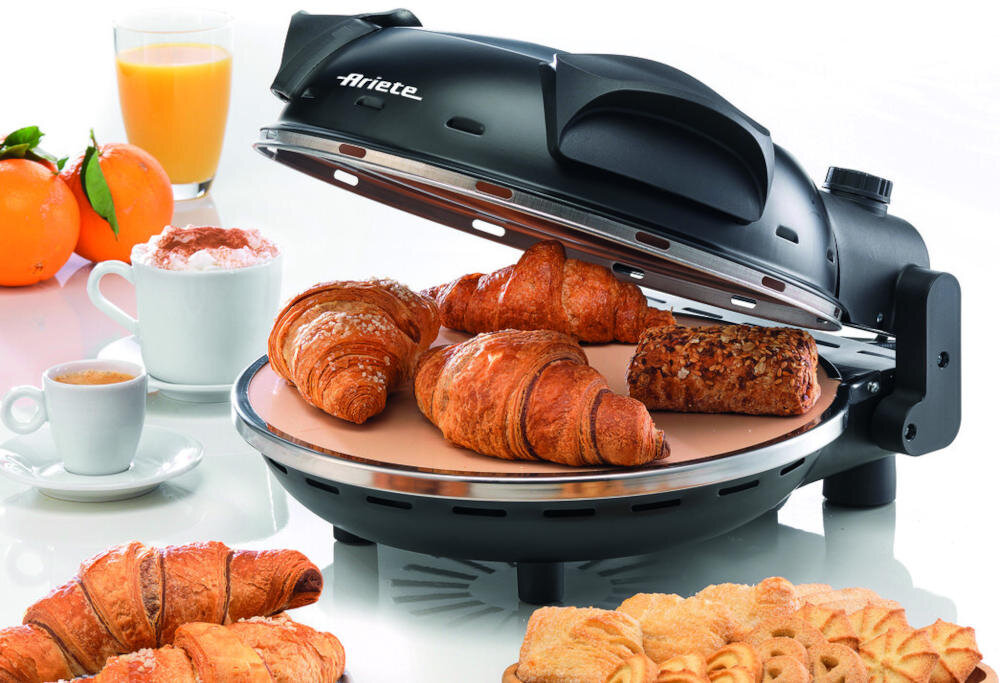 Na obrazie widoczny jest czarny piec do pizzy marki Ariete z otwartą pokrywą, na którym znajdują się croissanty i pieczywo. W tle widoczne są pomarańcze, filiżanka kawy i szklanka soku pomarańczowego.