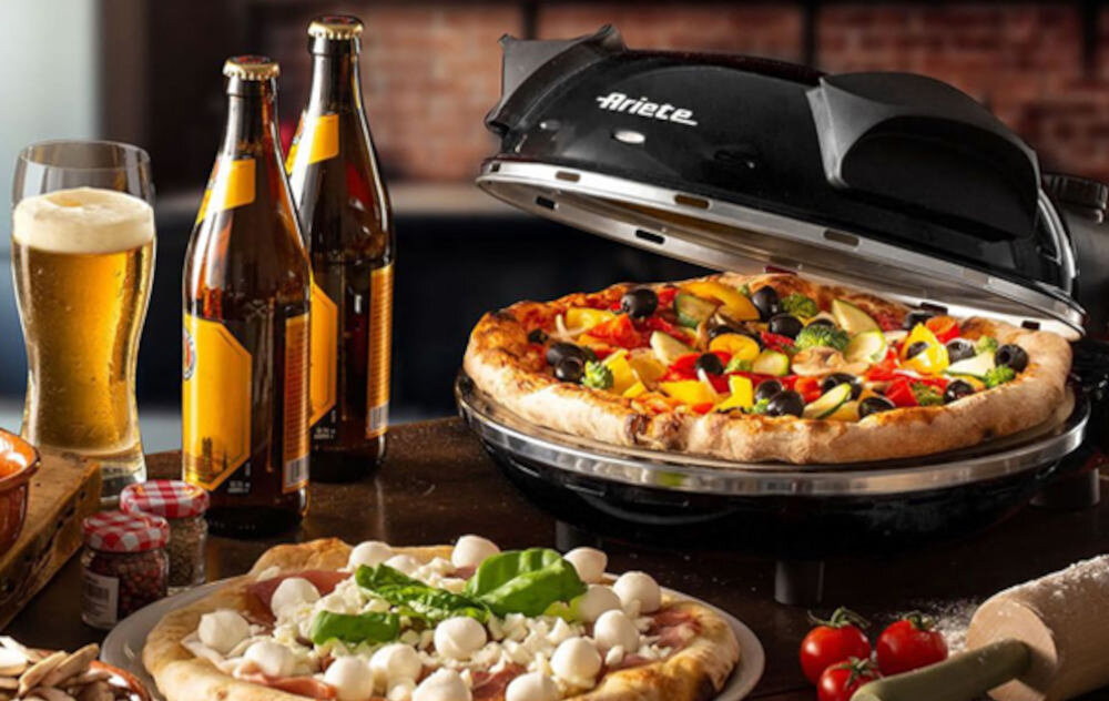 Na obrazie widać czarny piec do pizzy z logo \'Ariete\', w którym znajduje się pizza z różnymi dodatkami. Obok pieca stoją butelki piwa, szklanka z piwem oraz talerz z inną pizzą udekorowaną liśćmi bazylii i kulkami mozzarelli.