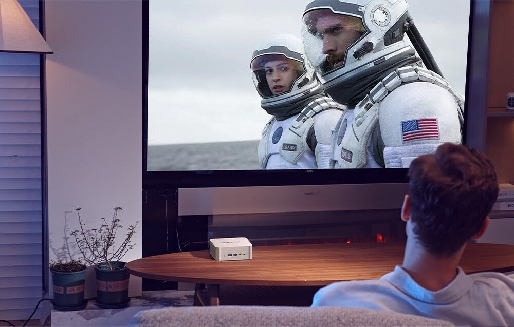 Komputer MINISFORUM X1-255 Mężczyzna ogląda film z astronautami na dużym telewizorze, a mały komputer stoi na szafce pod ekranem