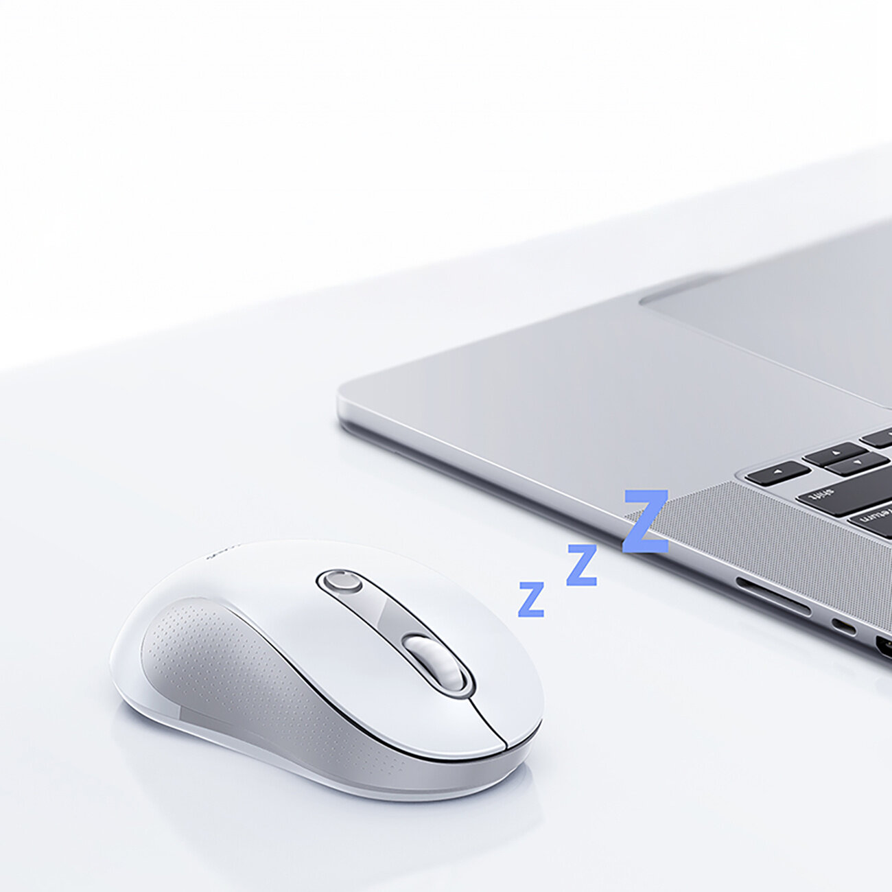 Na białym stole znajduje się biała mysz bezprzewodowa obok srebrnego laptopa. Obok myszy widoczny jest symbol \'zzz\' w kolorze niebieskim.