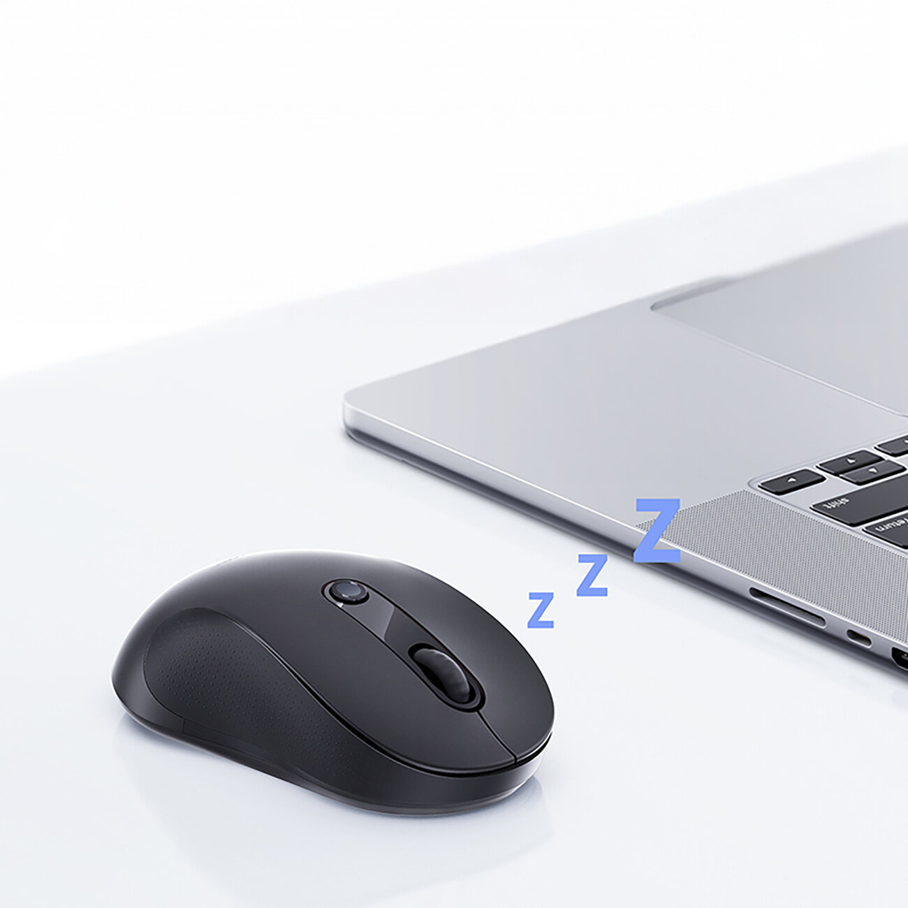 Na białym stole znajduje się czarna myszka bezprzewodowa obok srebrnego laptopa. Obok myszki widoczny jest symbol \'zzz\'.