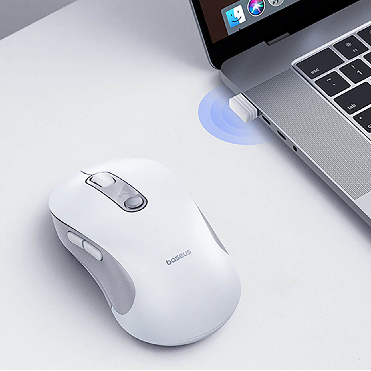 Na białym biurku znajduje się biała myszka komputerowa z napisem \'baseus\' obok laptopa z podłączonym odbiornikiem USB. Laptop ma widoczny fragment klawiatury i ekran z ikonami aplikacji.