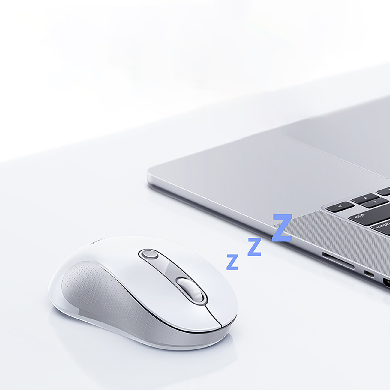 Na białym biurku znajduje się biała mysz bezprzewodowa obok srebrnego laptopa. Nad myszą widoczny jest niebieski symbol \'zzz\'.