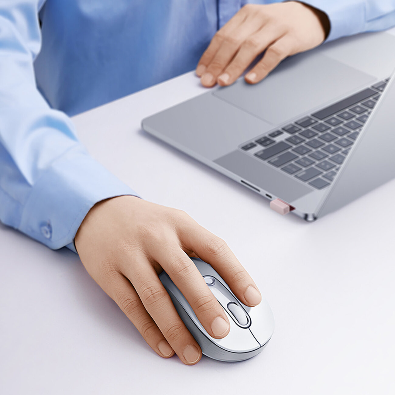 Osoba używa srebrnej, bezprzewodowej myszy komputerowej obok otwartego laptopa na białym biurku. Laptop ma widoczną klawiaturę i touchpad.