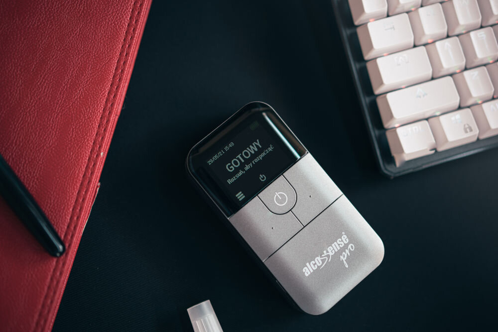 Na ciemnym tle znajduje się alkomat AlcoSense Pro z wyświetlaczem pokazującym tekst: \'GOTOWY Rozpocznij aby rozpocząć\'. Obok widoczna jest część klawiatury i czerwony notes.