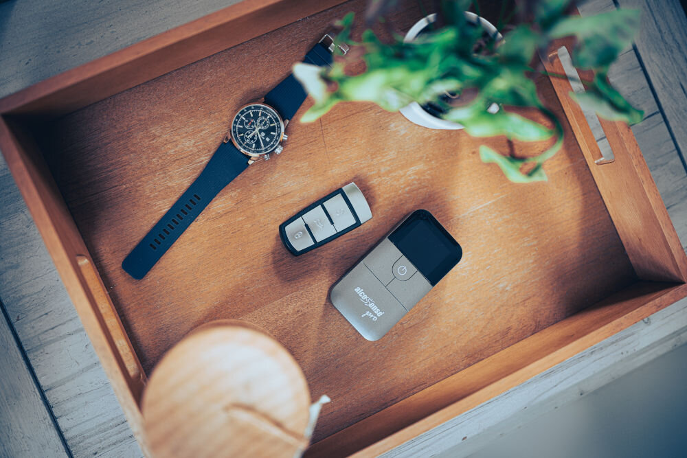 Na drewnianej tacy znajdują się zegarek z niebieskim paskiem, pilot z trzema przyciskami oraz urządzenie z napisem \'AlcoSense Pro\'. Obok widoczna jest zielona roślina w doniczce.