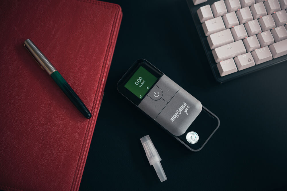 Na ciemnym biurku leży alkomat z wyświetlaczem pokazującym \'0.00 mg/l\', obok znajduje się ustnik, czerwony notes i długopis, a w tle klawiatura. Na alkomacie widoczne jest logo \'AlcoSense Pro\'.