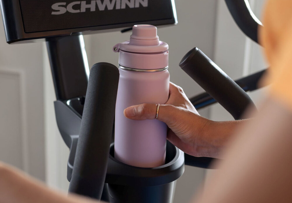 Orbitrek eliptyczny SCHWINN 590E 18kg 51cm osoba trzyma bidon w dłoni zbliżenie na uchwyt na bidon kółka transportowe solidna konstrukcja o wadze 96 kg obciążenie do 147 kg uchwyt na bidon