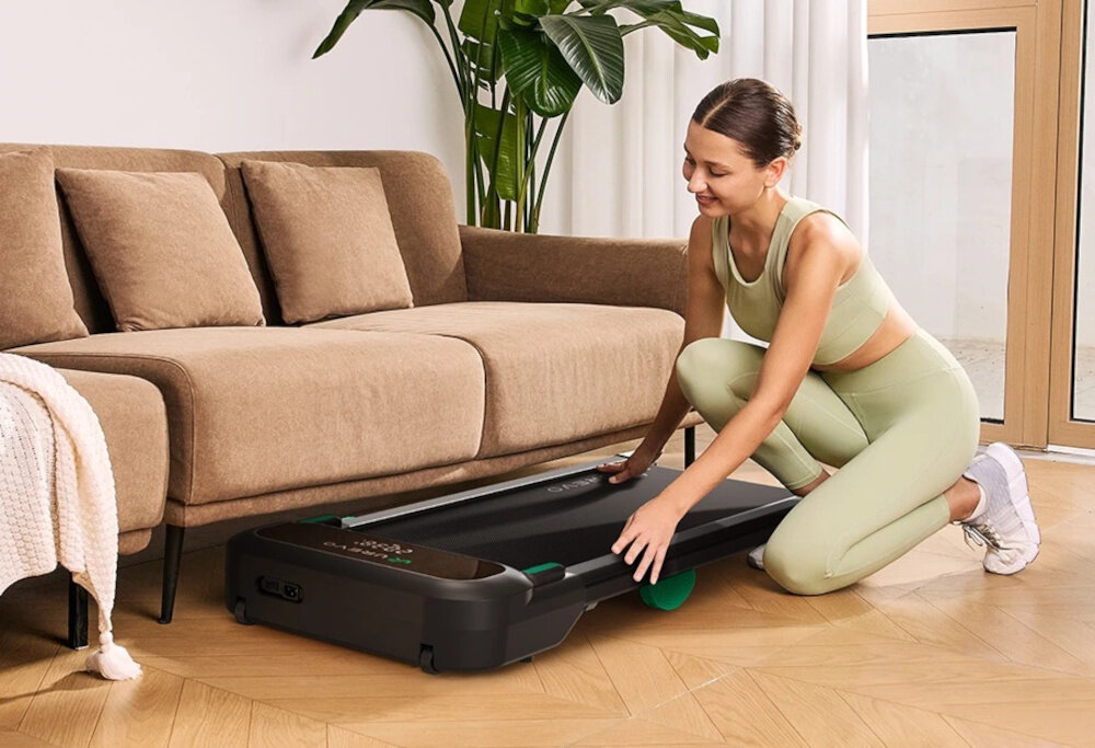 Bieżnia elektryczna UREVO Spacewalk 5L 600W 6 km-h pokój osoba chowa bieżnię pod kanapę w tle okno kwiat waga 23 kg wymiary 124 × 51,5 × 15,5 cm rolki transportowe nie zajmuje stałego miejsca nie ogranicza przestrzeni mieszkania