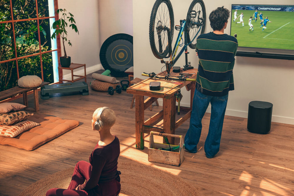 Soundbar JBL BAR 1000 MkII funkcja PureVoice 2.0 czyste dialogi optymalizacja głosu, osoba stojąca przed telewizorem oglądająca mecz, osoba siedzaca po turecku w słuchawkach na dywanie