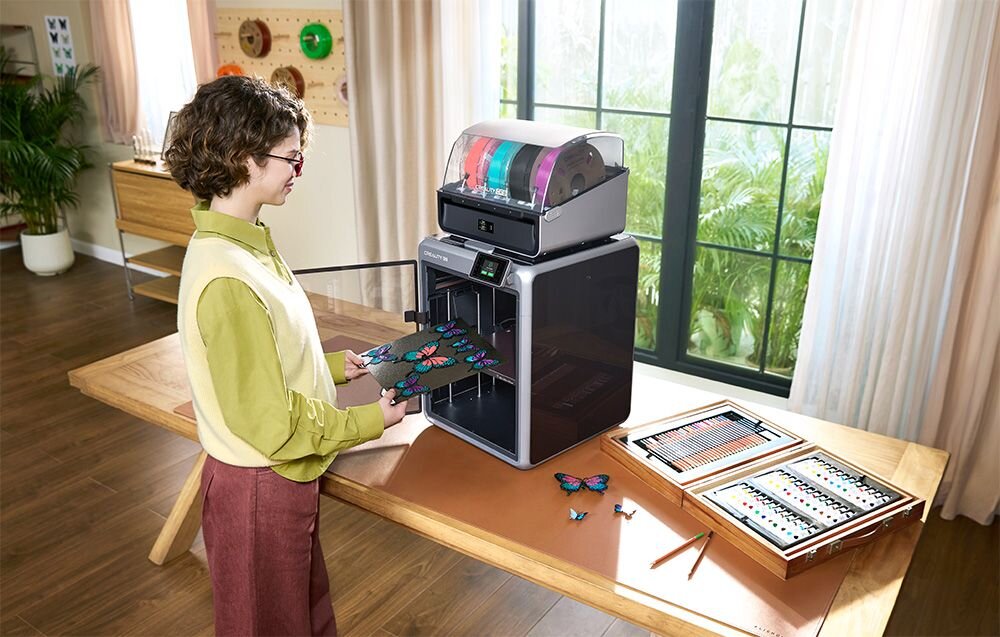 Kobieta wyjmująca platformę roboczą z wielokolorowymi wydrukami motyli z drukarki Creality K2 Combo zintegrowany system CFS do 16 kolorów niski poziom hałasu prędkość druku