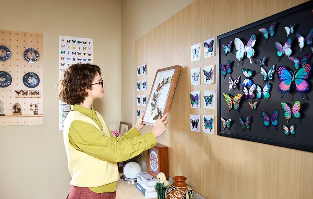 Kobieta wieszająca na ścianie ramkę z kolekcją motyli wydrukowanych w 3D w domowej pracowni elementy aluminiowe klasy lotniczej odporność na odkształcenia wymiary przestrzeń robocza wydajność