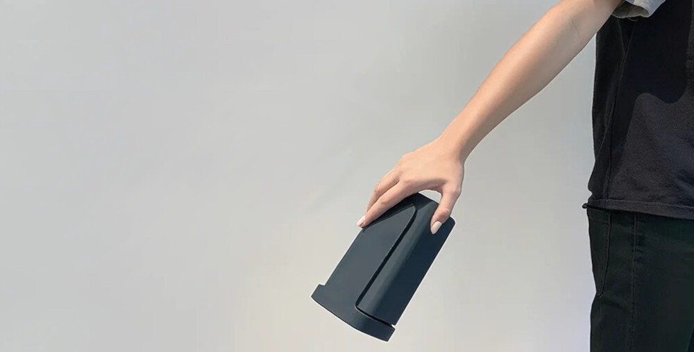 Osoba trzyma ciemny, stożkowaty przedmiot na tle jasnej ściany. Przedmiot ma gładką powierzchnię i prostą formę.