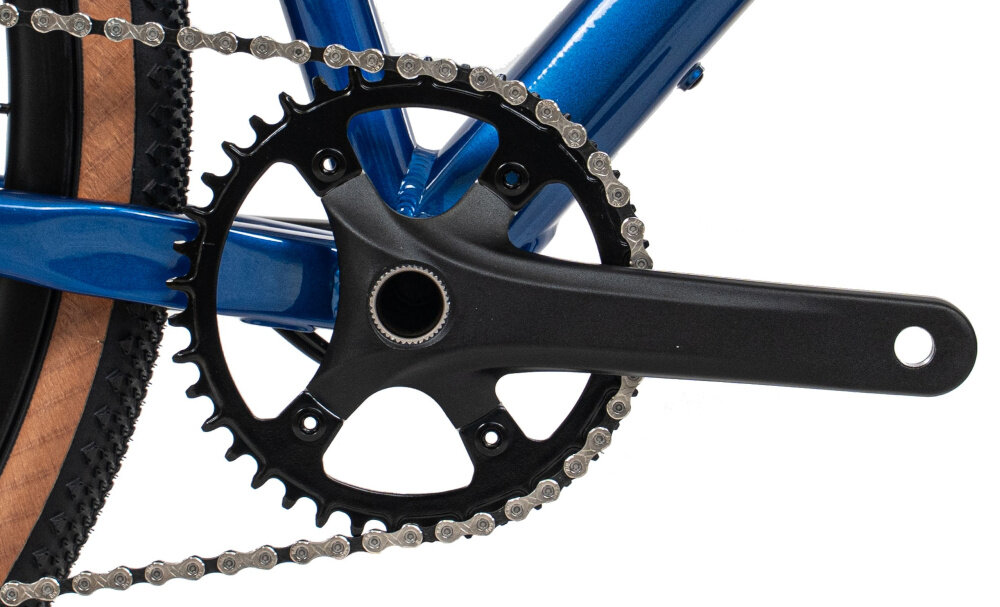 Zdjęcie przedstawia zbliżenie na mechanizm korbowy roweru z czarną zębatką i łańcuchem. Rama roweru jest w kolorze niebieskim, a opona ma brązowy bok.