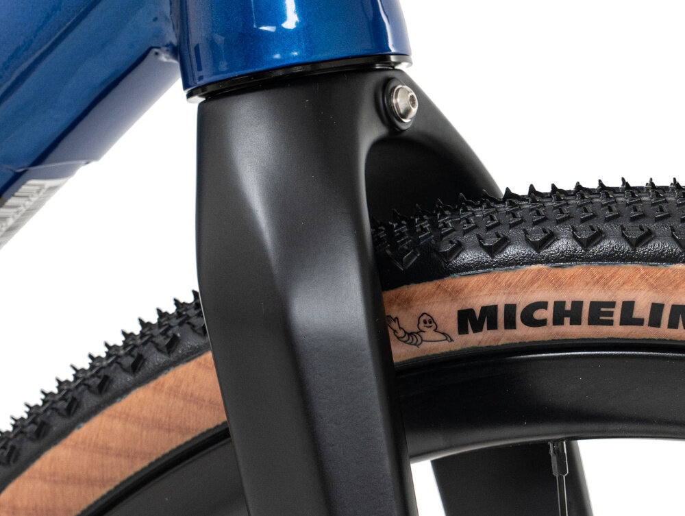Zbliżenie na przedni widelec roweru w kolorze niebieskim z czarnym wykończeniem oraz oponę z widocznym napisem \'MICHELIN\'.