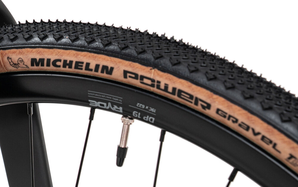 Rower gravel ROMET Aspre 2.1 U18 28 cali Różowy zbliżenie na przednie koło białe tło opony marki MICHELIN w rozmiarze 28 cali głęboki bieżnik skutecznie wspiera jazdę stabilny tor jazdy aluminiowe obręcze sztywność niska masa Piasty NOVATEC typu D981 z przodu w standardzie 12x100 mm i JOYTECH typu D982TSE w standardzie 12x142 mm z tyłu sztywność kół regularne sprawdzanie ciśnienia powietrza wartość na bocznej części opony