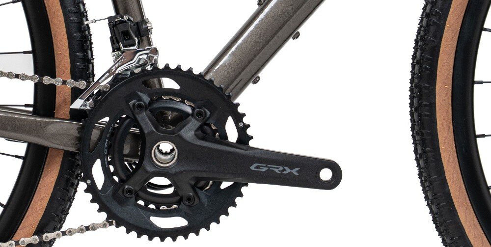 Zdjęcie przedstawia zbliżenie na mechanizm korbowy roweru z napisem \'GRX\'. Rama roweru ma grafitowy połysk, a opony mają jasnobrązowy bok.