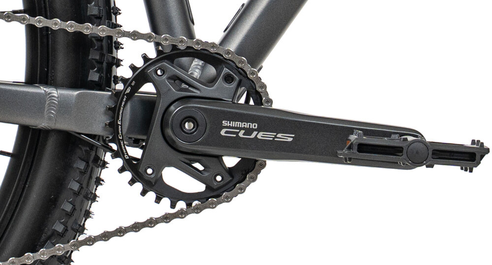 Zdjęcie przedstawia zbliżenie na mechanizm korbowy roweru z napisem \'Shimano Cues\'. Widoczny jest łańcuch oraz część tylnego koła z oponą.