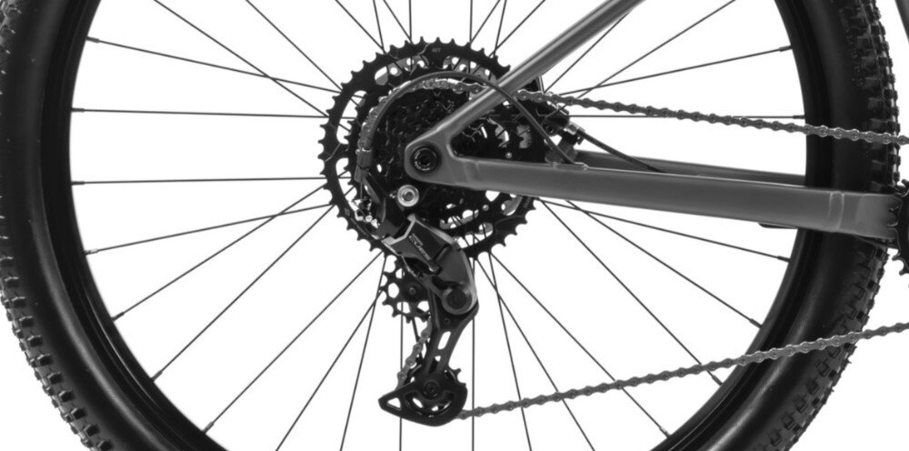 Zdjęcie przedstawia tylne koło roweru z widocznym mechanizmem przerzutki i zębatkami. Opona ma wyraźny bieżnik, a rama roweru jest w kolorze antracytowym.