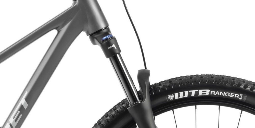 Zbliżenie na przednią część roweru górskiego z widocznym amortyzatorem i oponą z napisem \'WTB RANGER 2.4\'. Rama roweru jest w kolorze antracytowym z widocznym fragmentem napisu \'ROMET\'.