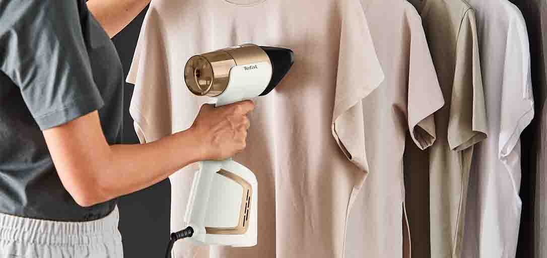 Kobieta trzyma w ręku parownicę DT9820 AeroSteam i przykłada ją do koszulki na wieszaku, w tle więcej koszulek.