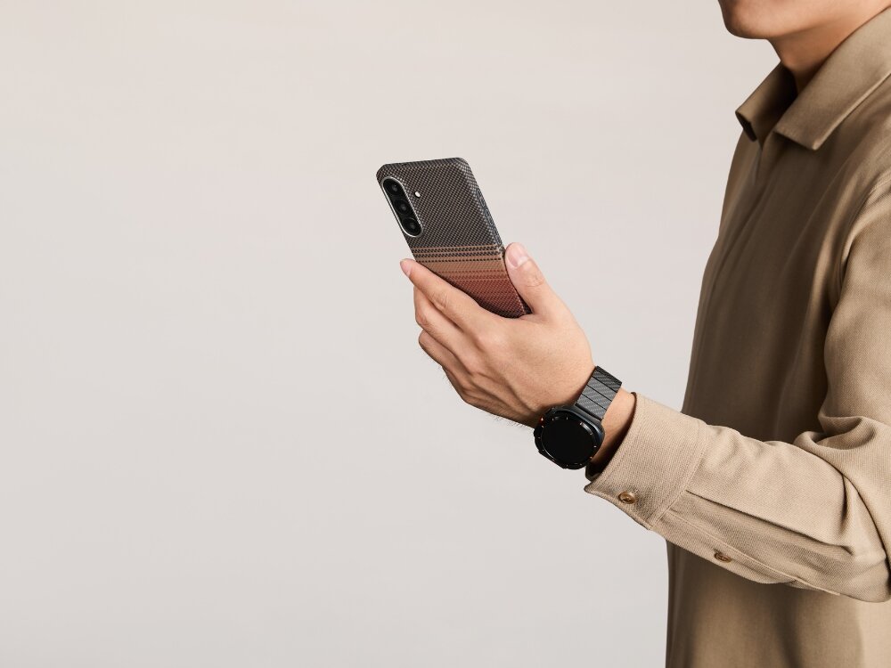 Etui PITAKA Ultra-Slim MagSafe Samsung Galaxy S26 - włókno aramidowe, smukłe, MagSafe Smartfon Mężczyzna trzymający smartfon w etui Pitaka z serii Sunset o wzorze aramidowym, prezentujący dopasowany pasek do zegarka typu smartwatch.