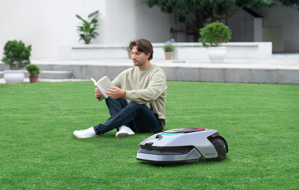 Robot koszący DREAME A2 robot na trawie w tle osoba siedząca na trawniku czyta książkę dalej dom taras i kwiaty zaawansowany silnik piastowy praca na terenach o nachyleniu do 50% 26,5° poziom hałasu 55 dB