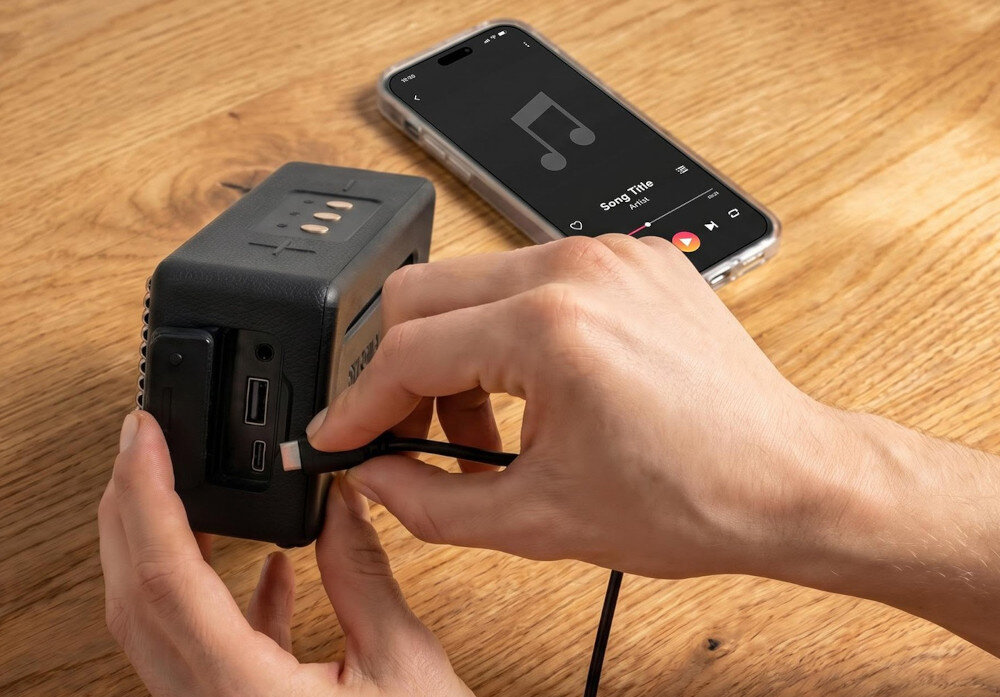 Osoba podłącza kabel do czarnego głośnika mobilnego z widocznymi portami USB. Obok leży smartfon z otwartą aplikacją muzyczną, na ekranie widoczny jest symbol nuty i tekst \'Song Title\'.