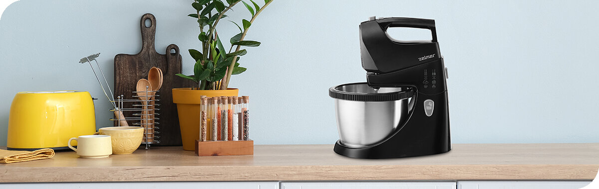 Na blacie kuchennym stoi czarny mikser z misą marki Zelmer. Obok znajdują się różne akcesoria kuchenne, w tym żółty czajnik i drewniane deski do krojenia.