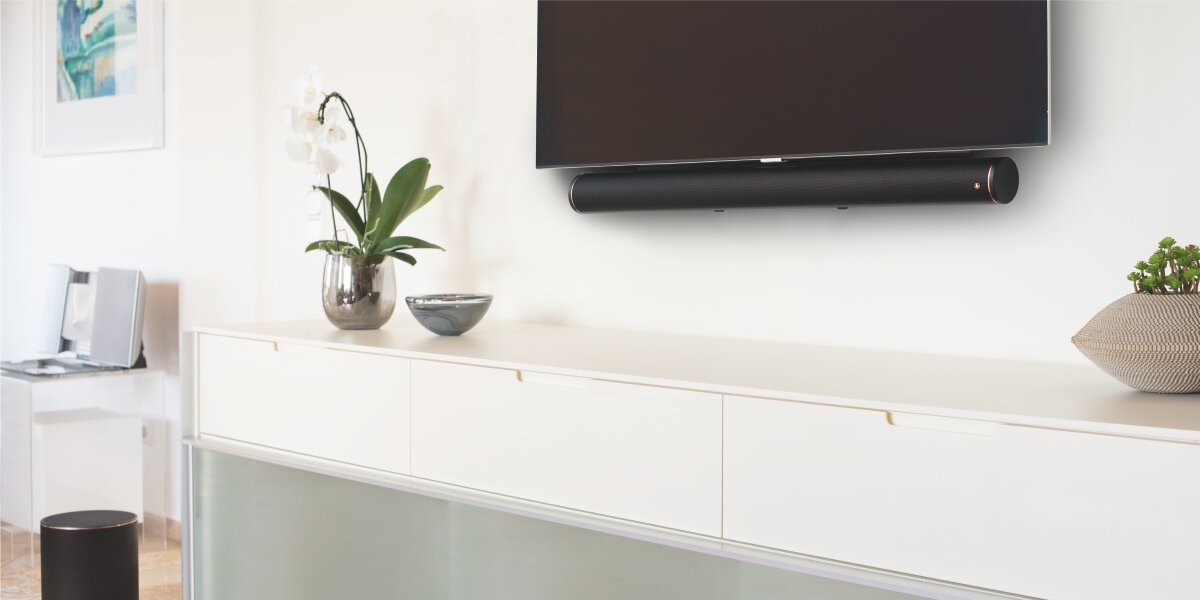 Uchwyt do soundbara HAMA 220883 soundbar zamontowany dyskretnie pod telewizorem na ścianie w jasnym, nowoczesnym salonie. Pod urządzeniem znajduje się biała komoda z dekoracjami.