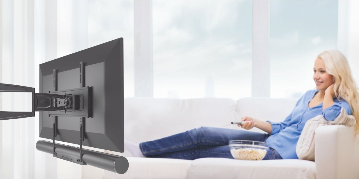 Uchwyt do soundbara HAMA 220884 Kobieta siedząca na kanapie z pilotem i miską popcornu, oglądająca telewizor z zamontowanym pod nim soundbarem. 