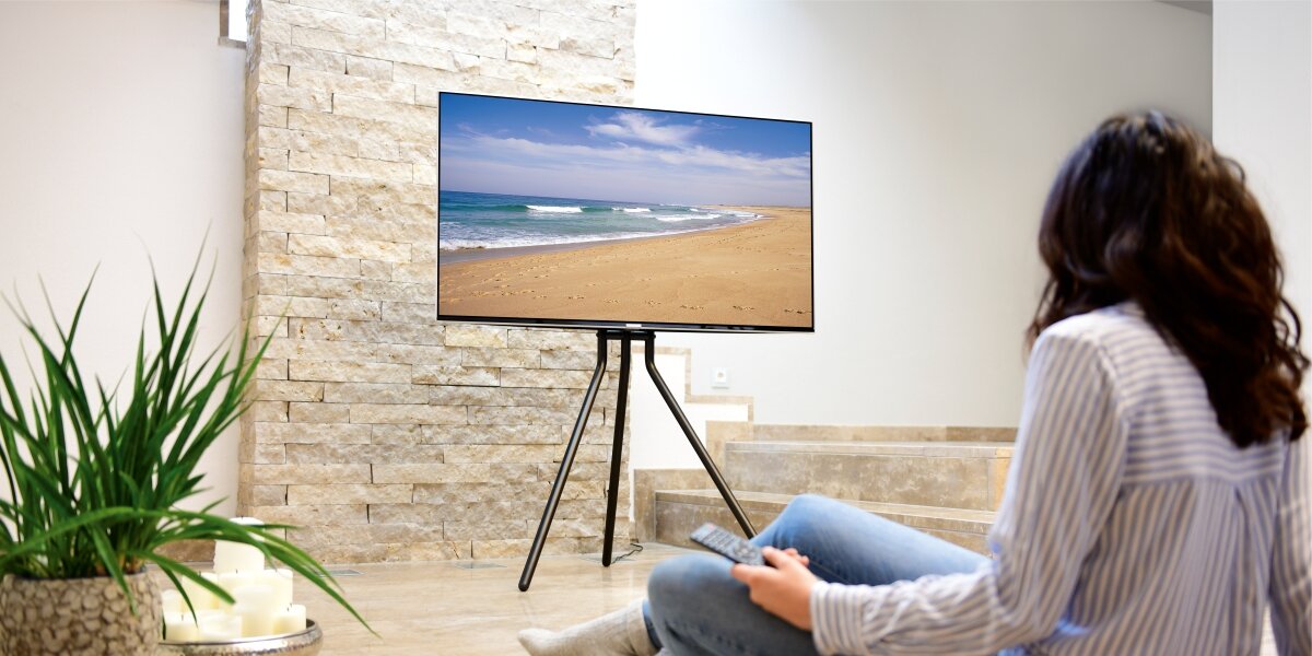 Stojak podłogowy HAMA do TV 37-75 cali 220870 Czarny Kobieta siedząca na podłodze ogląda plażę na telewizorze zamontowanym na eleganckim, czarnym stojaku typu tripod.