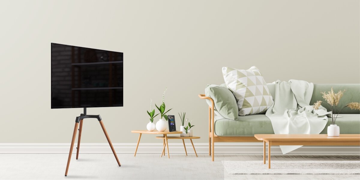 Stojak podłogowy HAMA do TV 32-65 cali 220873 Designerski stojak pod telewizor typu trójnóg z drewnianymi nogami w jasnym salonie z zieloną kanapą i nowoczesnymi stolikami.