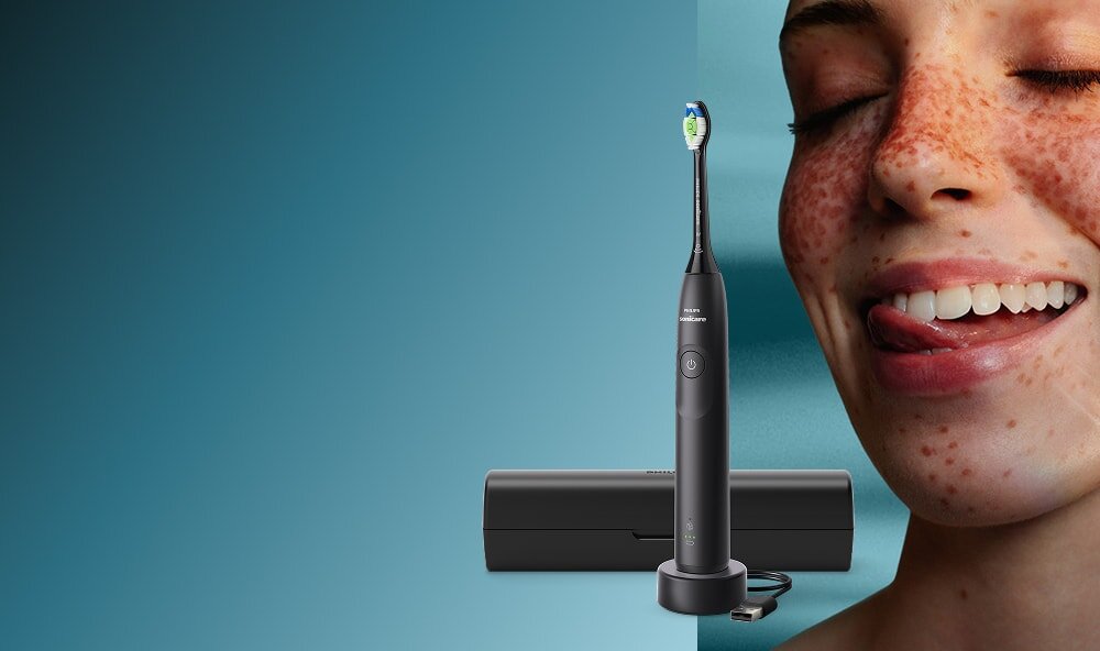 Zbliżenie na twarz uśmiechniętej kobiety, która językiem przesuwa po zębach. Obok szczoteczka soniczna z etui.