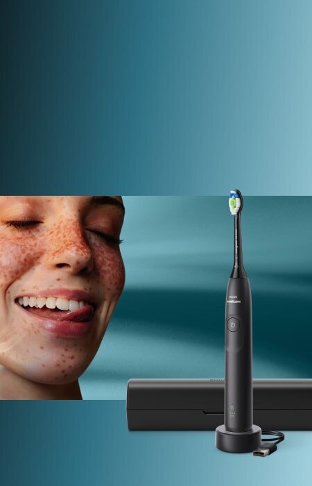 Zbliżenie na twarz uśmiechniętej kobiety, która językiem przesuwa po zębach. Obok szczoteczka soniczna z etui.