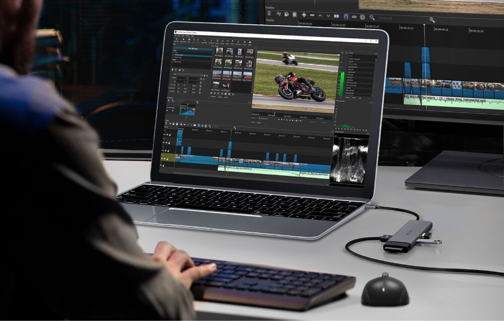 Hub TP-LINK UH7021C Mężczyzna siedzi przy biurku i pracuje na laptopie z podłączonym wieloportowym adapterem, do którego wpięte są pendrive'y oraz kable, a w tle widoczny jest drugi monitor z włączonym programem do edycji wideo
