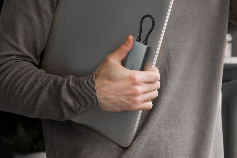 Hub TP-LINK UH7021C Mężczyzna trzyma w jednej dłoni zamkniętego laptopa oraz szary adapter z kablem schowanym w specjalnym uchwycie, co podkreśla mobilność urządzenia
