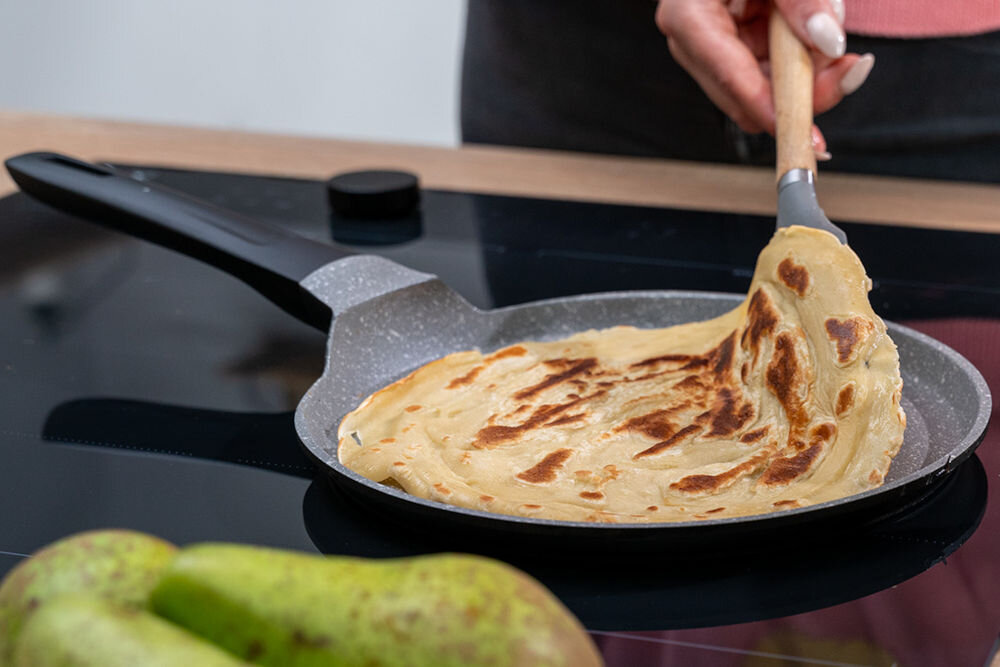 Na obrazie widać patelnię z naleśnikiem, który jest przewracany za pomocą drewnianej łopatki. W tle widoczne są dwie gruszki na blacie kuchennym.