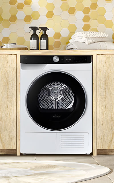 Suszarka DV90DG6845LKU4 w jasnym pomieszczeniu Biała suszarka Samsung w zabudowie z jasnego drewna, na tle żółto-białych płytek, dostępna w Media Expert.