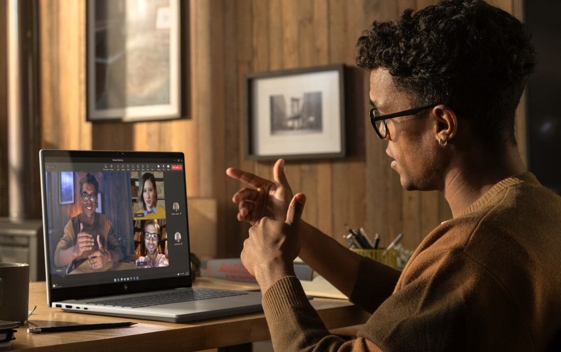 Mężczyzna w brązowym swetrze i okularach gestykulujący podczas wideokonferencji na laptopie HP OmniBook, na ekranie widoczni czterej uczestnicy spotkania online, w tle nowoczesne, przytulne wnętrze biurowe.  