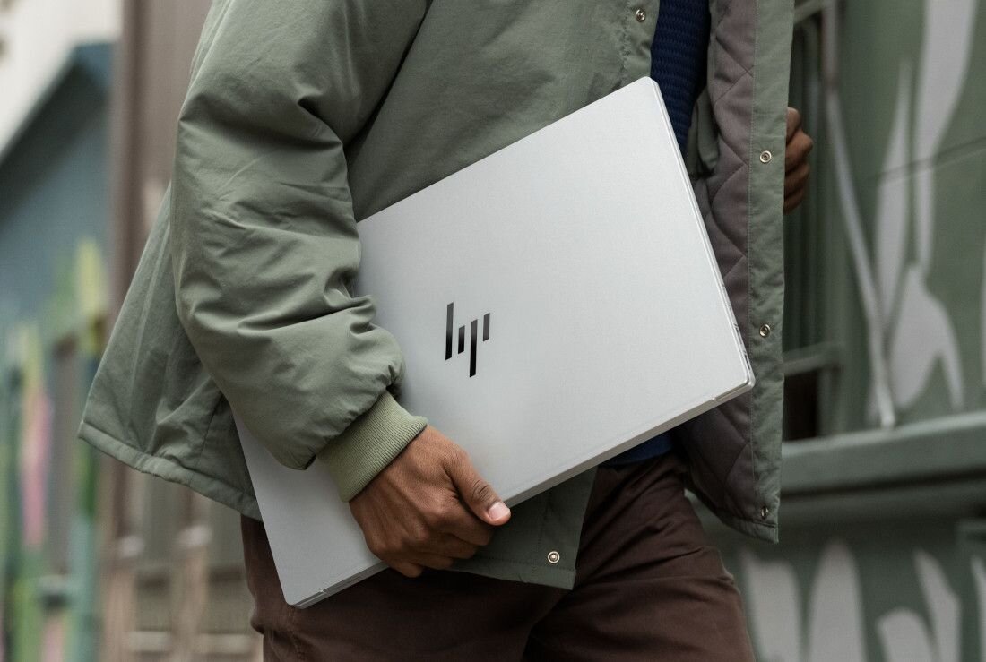 Mężczyzna w zielonej kurtce trzymający zamknięty, smukły laptop HP OmniBook w srebrnej obudowie, idący ulicą, podkreślający mobilność, wytrzymałość i elegancki design urządzenia w miejskim otoczeniu. - Wytrzymałość 