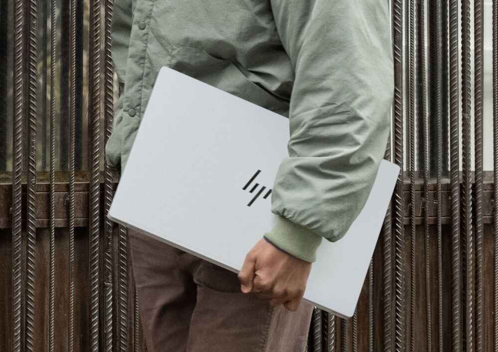 Mężczyzna w zielonej kurtce trzymający zamknięty laptop HP OmniBook w srebrnej obudowie, idący wzdłuż metalowego ogrodzenia, podkreślający mobilność i smukły design urządzenia w miejskim otoczeniu. - Opis serii OmniBook 7 NGAI 