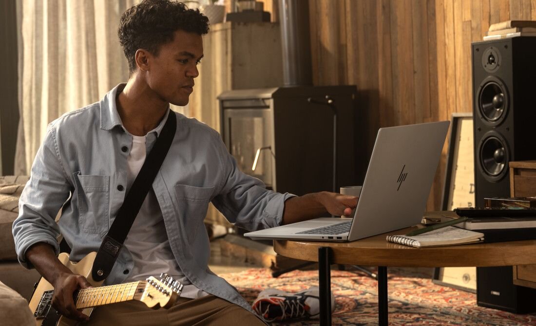 Młody mężczyzna z gitarą elektryczną na ramieniu korzystający z laptopa HP OmniBook w domowym salonie, obok głośniki i kominek, atmosfera kreatywnej pracy i rozrywki. - Pamięć LPDDR5x   